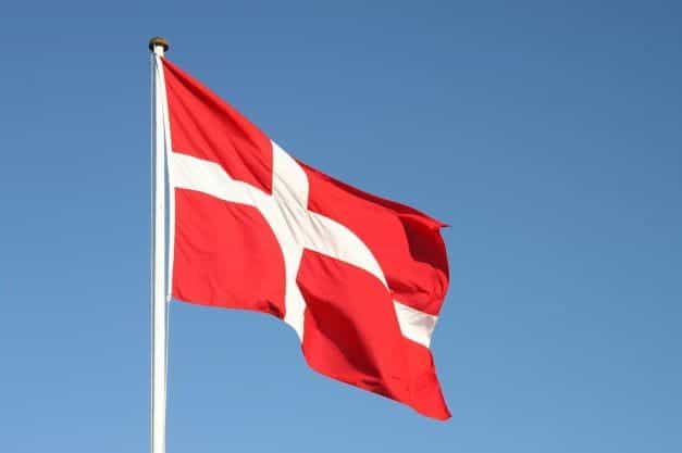 The Danish flag, white cross on red background, on a flagpole with blue sky.