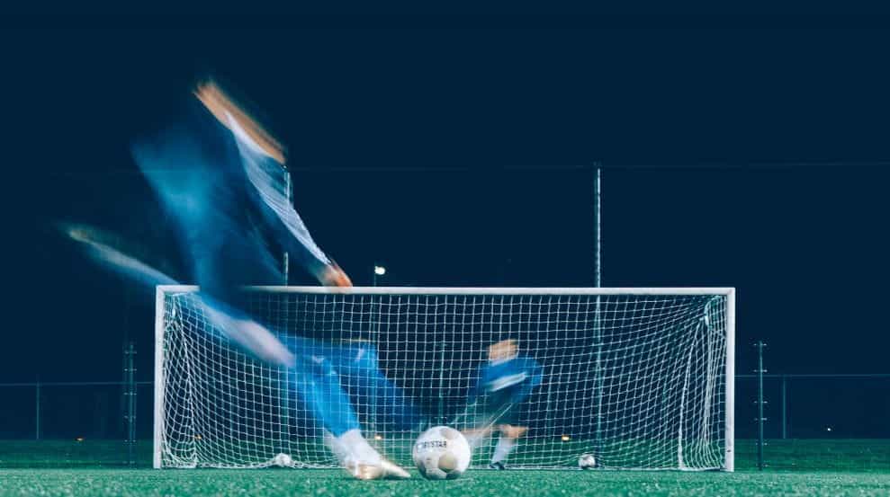 A time lapse image of two soccer players in blue uniforms kicking and defending a ball in front of a goal.