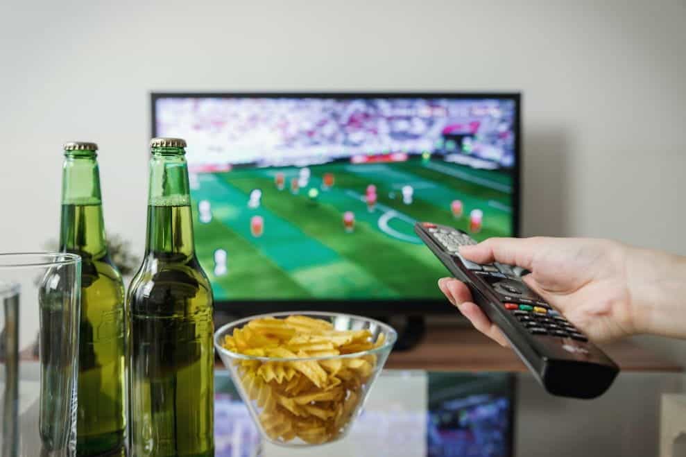 A hand holding a remote pointing at a TV showing a football match, with beer and crisps.