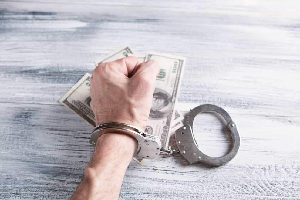 A first in handcuffs sits on top of spread out banknotes on a table.
