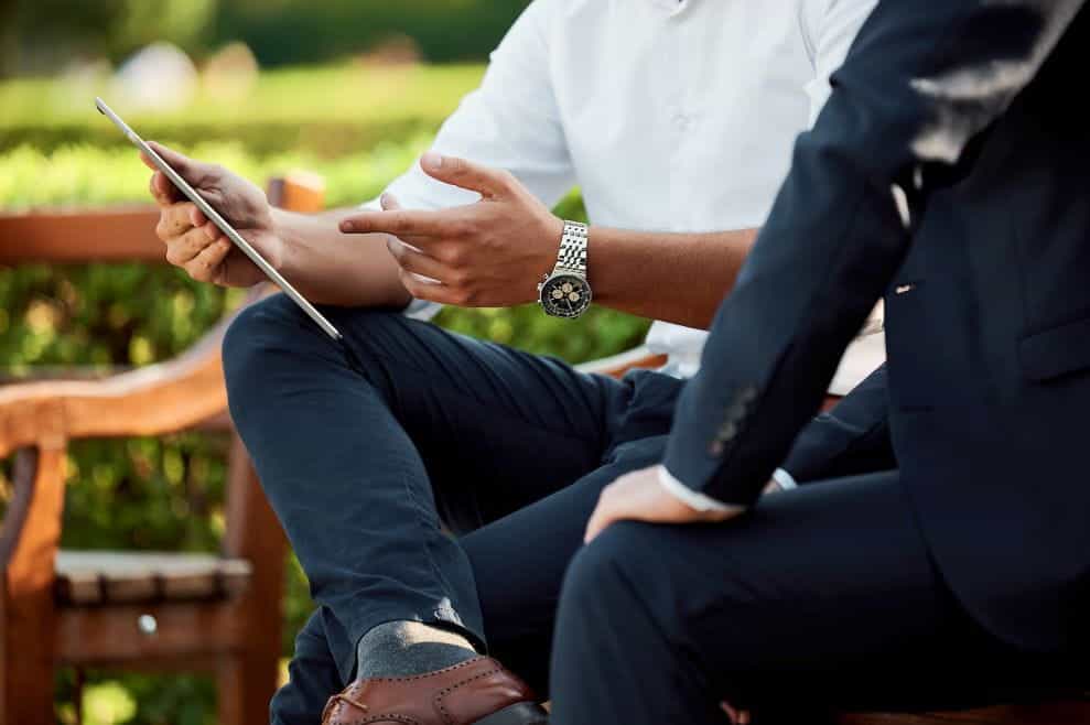 Two men in formal clothing sitting discuss over an iPad in a park.