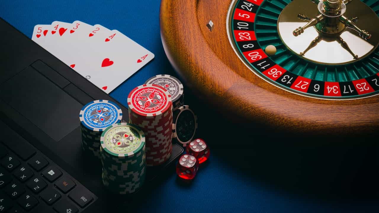 A roulette wheel, poker chips, dice and playing cards next to a laptop.