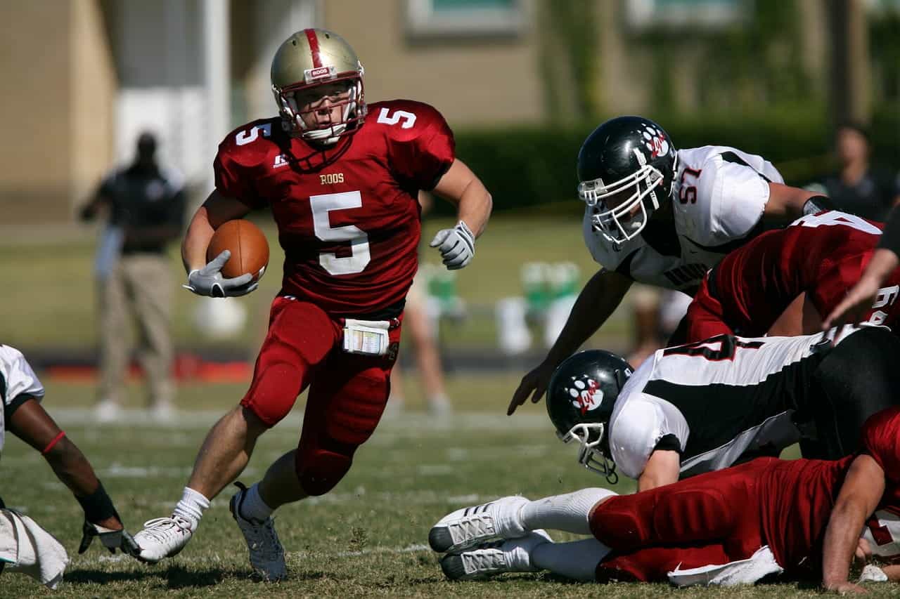 A male American football player running with the ball and executing a maneuver by avoiding rival players during an outdoor game on a football field.