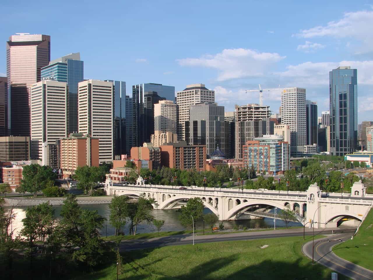 The downtown skyline of Calgary, Alberta, Canada, featuring several skyscrapers and tall buildings, construction cranes, a bridge going over a river, and a small park on the river embankment.