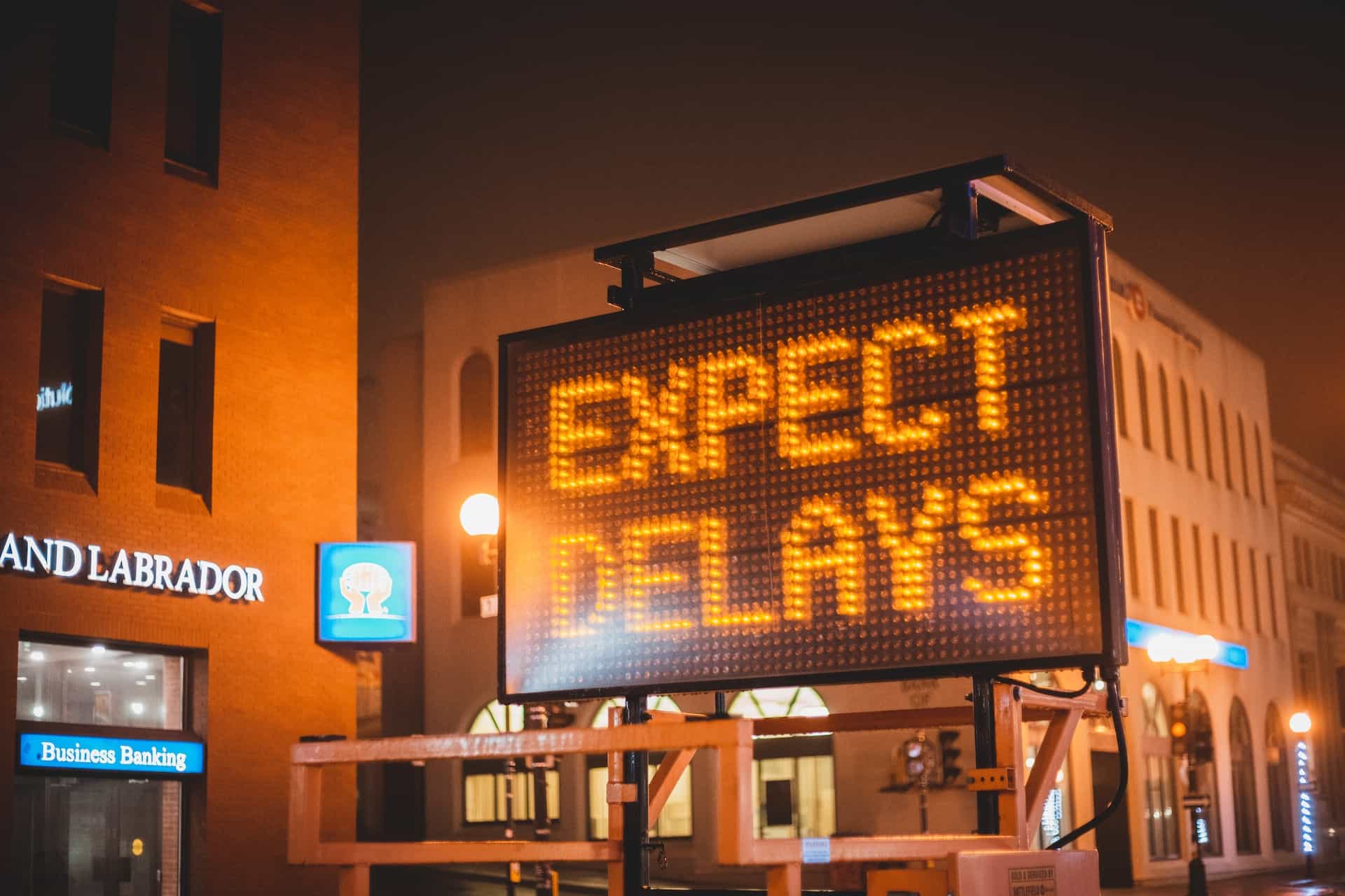 A traffic sign in a street at night reads EXPECT DELAYS in orange lights.